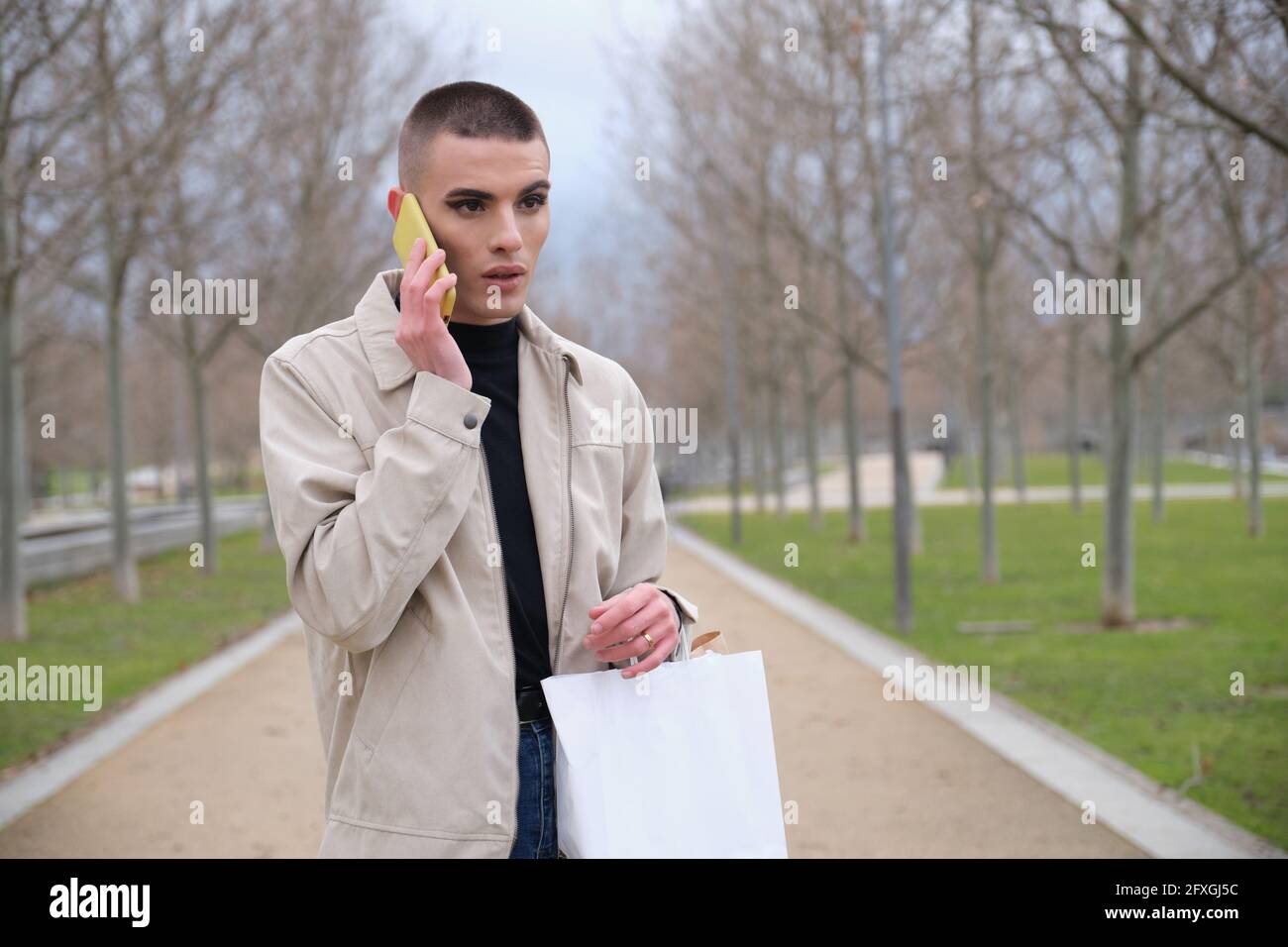 Jeune homme portant un maquillage tenant des sacs de shopping et parlant sur un smartphone dans un parc. Queer personne. Banque D'Images
