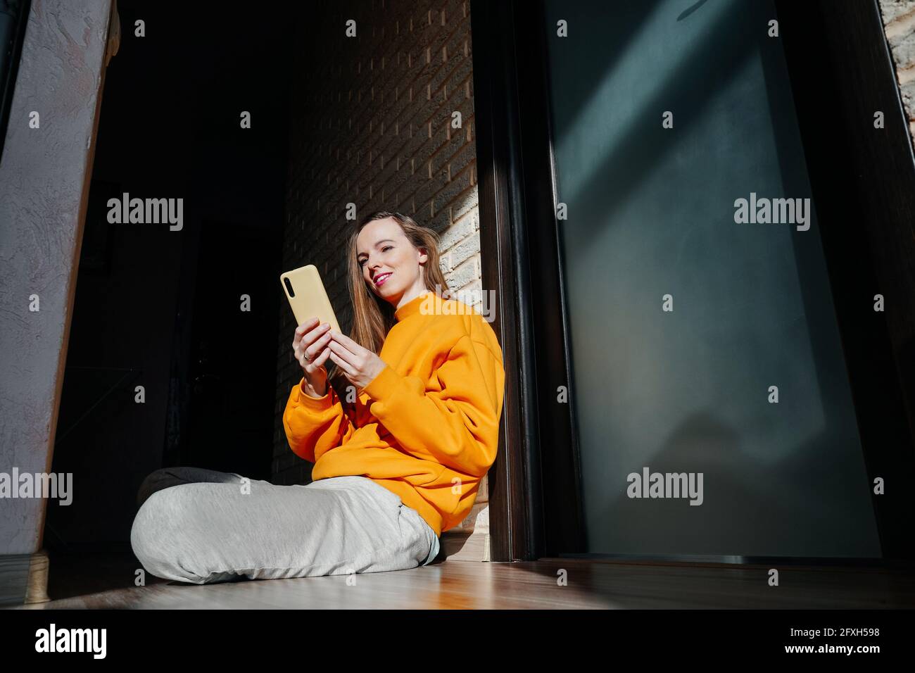Une jeune femme travaille avec un téléphone portable à la maison. Sweat-shirt orange jaune vif et pantalon gris. Banque D'Images