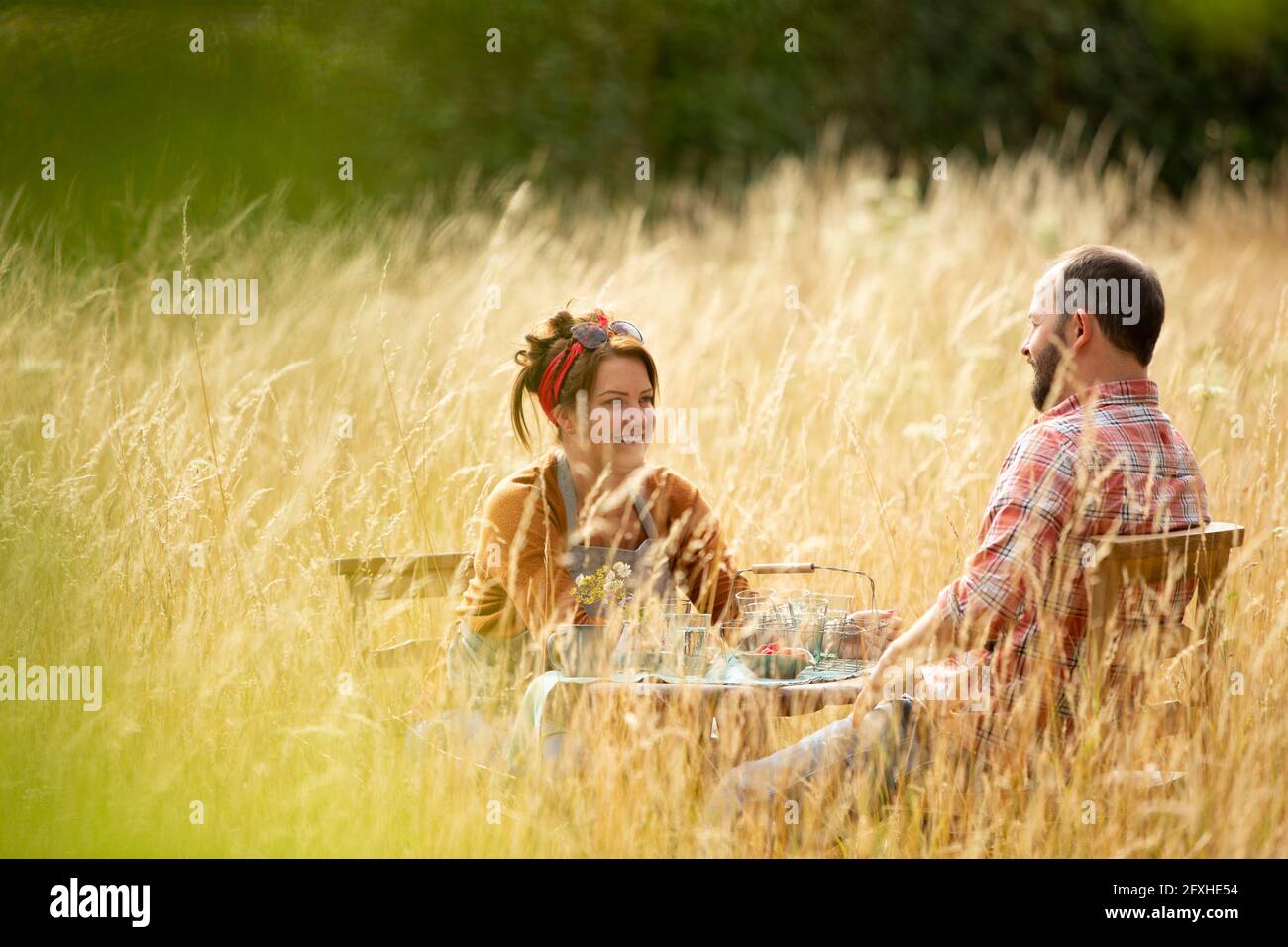 Couple heureux de se détendre à table dans l'herbe haute ensoleillée Banque D'Images Couple heureux de se détendre à table dans l'herbe haute ensoleillée Banque D'Images