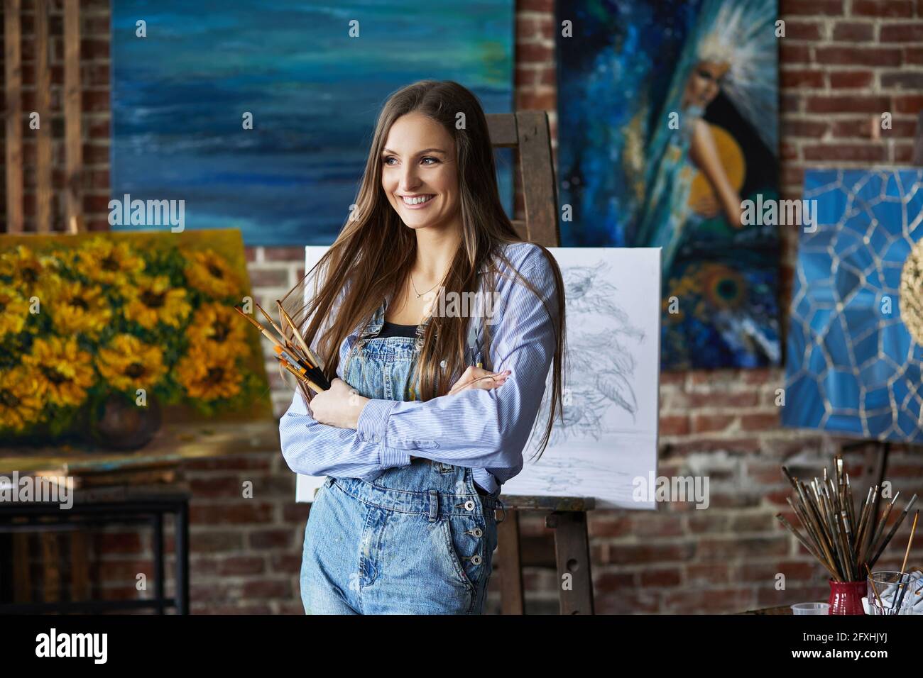 Portrait d'une jeune artiste souriante avec ses œuvres en studio d'art. Peintre tenant des pinceaux d'art. Processus créatif, détente, loisir, passe-temps Banque D'Images