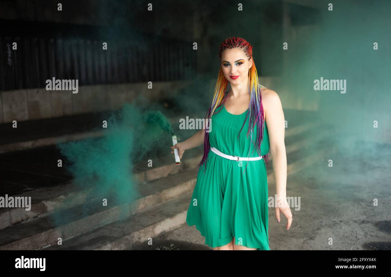 Jolie fille dans une robe verte avec des tresses africaines et maquillage  coloré posé avec de la fumée vert vif dans les rues d'une ville de  printemps chaude Photo Stock - Alamy