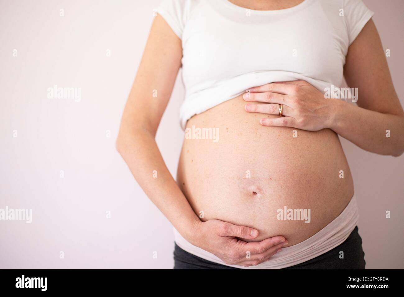Femme enceinte portant un t-shirt blanc avec les mains sur le ventre sur fond blanc. Vue centrale. Arrière-plan avec espace de copie. Banque D'Images