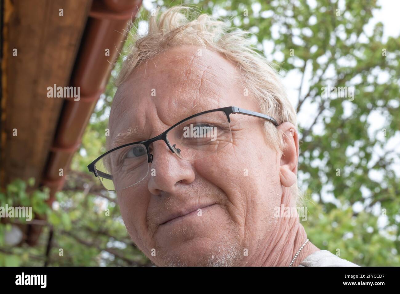 Un adulte mature d'apparence européenne aux cheveux blonds, portant des lunettes, regarde la caméra et sourit, dans la rue sur un fond de gre Banque D'Images