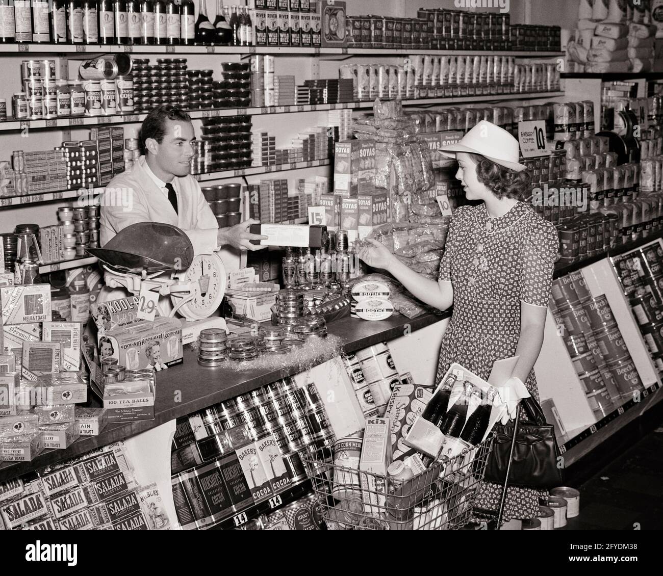 DANS LES ANNÉES 1940, UNE FEMME FAIT SES COURSES DANS UN MAGASIN D'ALIMENTATION POUSSANT LE CHARIOT À L'ACCEPTER UN PAQUET DE CLERK HOMME DEBOUT DERRIÈRE LE COMPTOIR - S7569 HAR001 HARS 1 STYLE DE VIE COMMIS FEMMES EMPLOIS COPIE ESPACE DEMI-LONGUEUR FEMMES PERSONNES MAGASINS MÂLES FORFAIT B&W SHOPPER SHOPPERS OCCUPATION VENDRE DES MÉTIERS DE L'ÉPICIER DE SERVICE À LA CLIENTÈLE LES MAGASINS ACCEPTANT LE COMMERCE ÉLÉGANT MOYEN-ADULTE HOMME MOYEN-ADULTE FEMME MOYEN-ADULTE COMMERCIAL NOIR ET BLANC ENTREPRISES CAUCASIENNES ETHNICITÉ HAR001 À L'ANCIENNE Banque D'Images