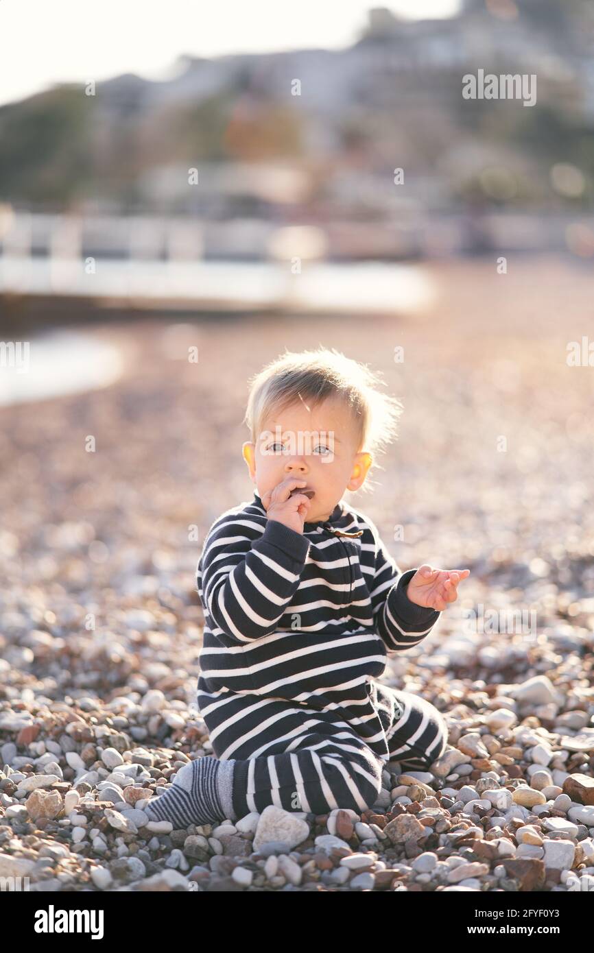 Un petit garçon en combinaison rayée est assis sur ses genoux une plage de  galets avec un galet dans sa bouche Photo Stock - Alamy