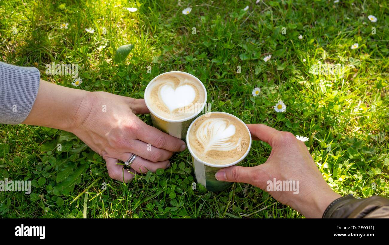 Deux mains de deux femmes tenant un à emporter recyclage café cappuccino tasses avec coeur mousse de lait forme décoration sur fond vert prairie. Banque D'Images