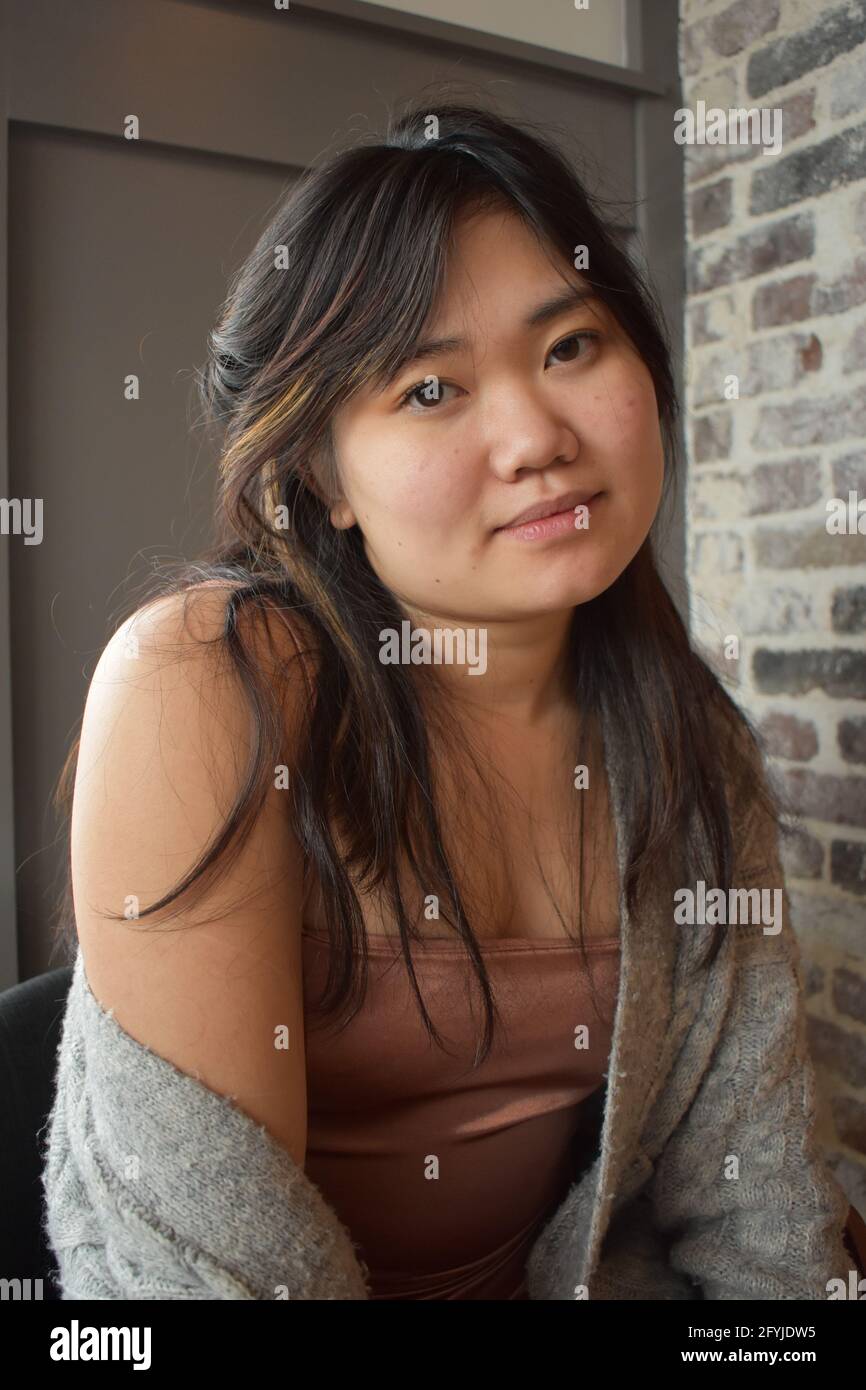 Modèle asiatique américain posant pour la caméra, vêtu d'une robe courte et d'un chandail, jambes croisées, main sur le menton et sourire à la caméra Banque D'Images