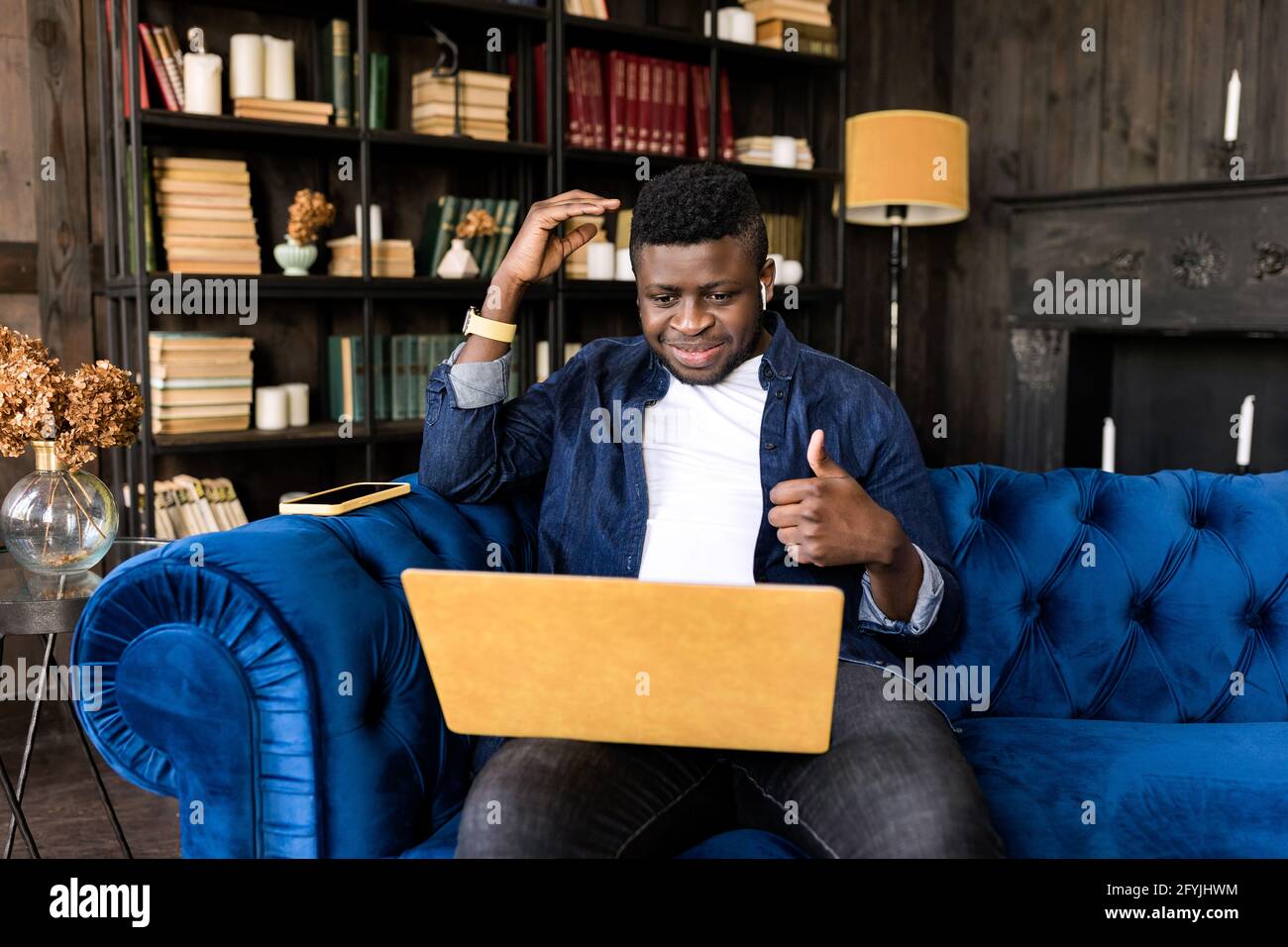 Jeune homme d'affaires élégant assis sur un canapé, montrant le pouce jusqu'à la vidéoconférence en ligne avec un ordinateur portable jaune pour des collègues. Indépendant ou Banque D'Images