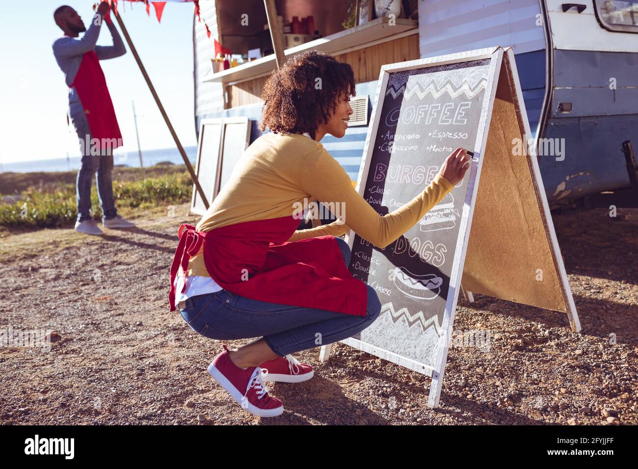 Divers couple ouvrant et préparant un camion de nourriture en bord de mer le jour ensoleillé, femme écrivant sur le tableau de menu Banque D'Images