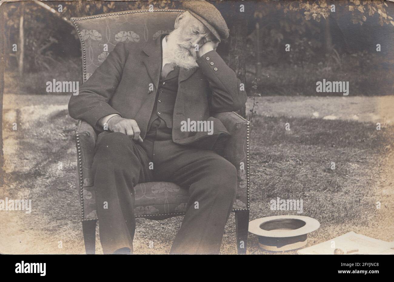 Carte postale photographique vintage du début du XXe siècle montrant un homme âgé avec une barbe blanche et portant un costume et une casquette en tissu snoozing à l'extérieur dans sa chaise. Banque D'Images