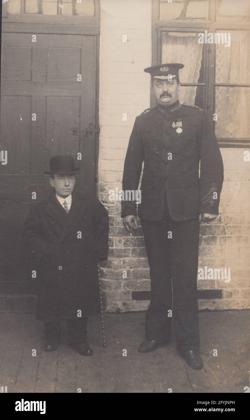 Carte postale photographique britannique vintage du début du XXe siècle montrant un homme en uniforme portant une médaille et un homme avec le nanisme. Banque D'Images