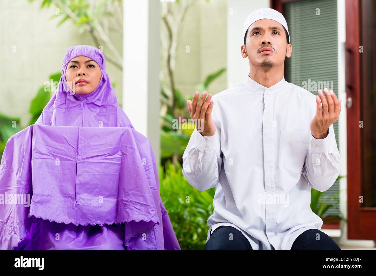 Couple musulman d'Asie, l'homme et de la femme, en priant à la maison assis sur les tapis de prière dans leur maison devant le jardin tropical Banque D'Images