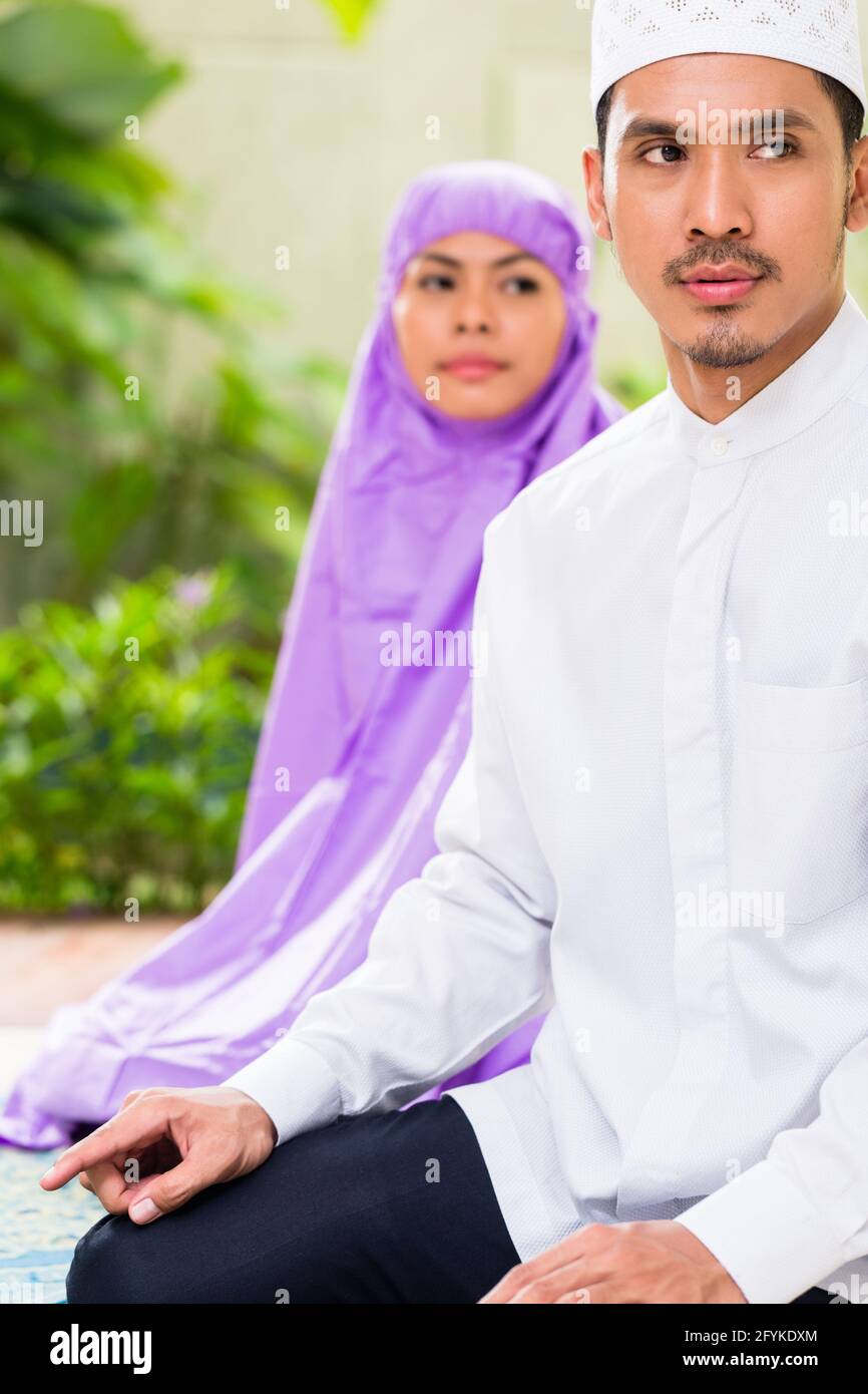 Couple musulman d'Asie, l'homme et de la femme, en priant à la maison assis sur les tapis de prière dans leur maison devant le jardin tropical Banque D'Images