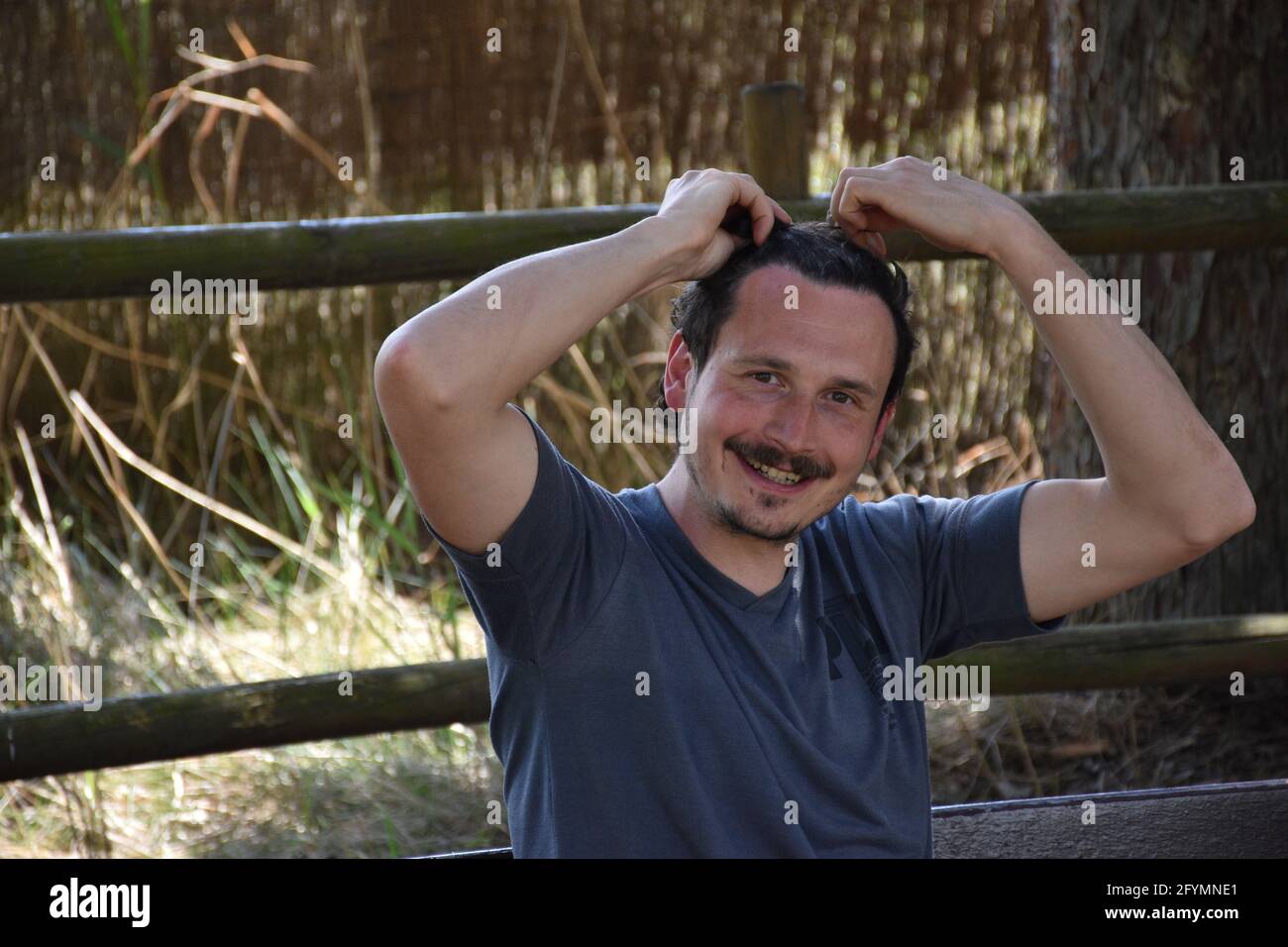Homme souriant avec moustache et bouc, bras levés faisant une queue de cheval dans ses cheveux. Zone ombragée avec lits en roseau en arrière-plan. Banque D'Images