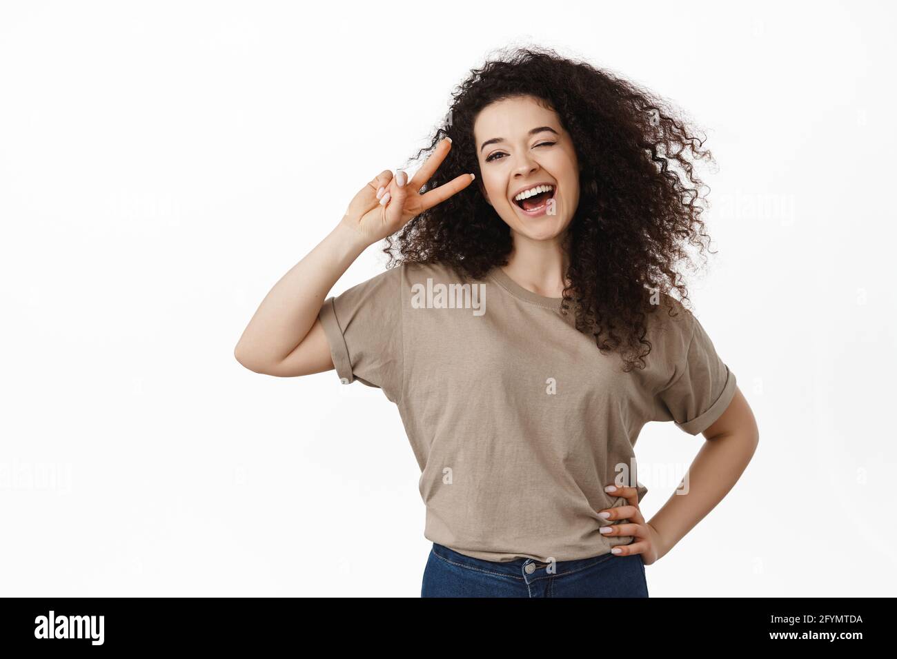 Portrait d'une femme brune heureuse et positive, riant et souriant, montrant le signe de la paix v près de l'œil, pose de kawaii, debout sur fond blanc Banque D'Images
