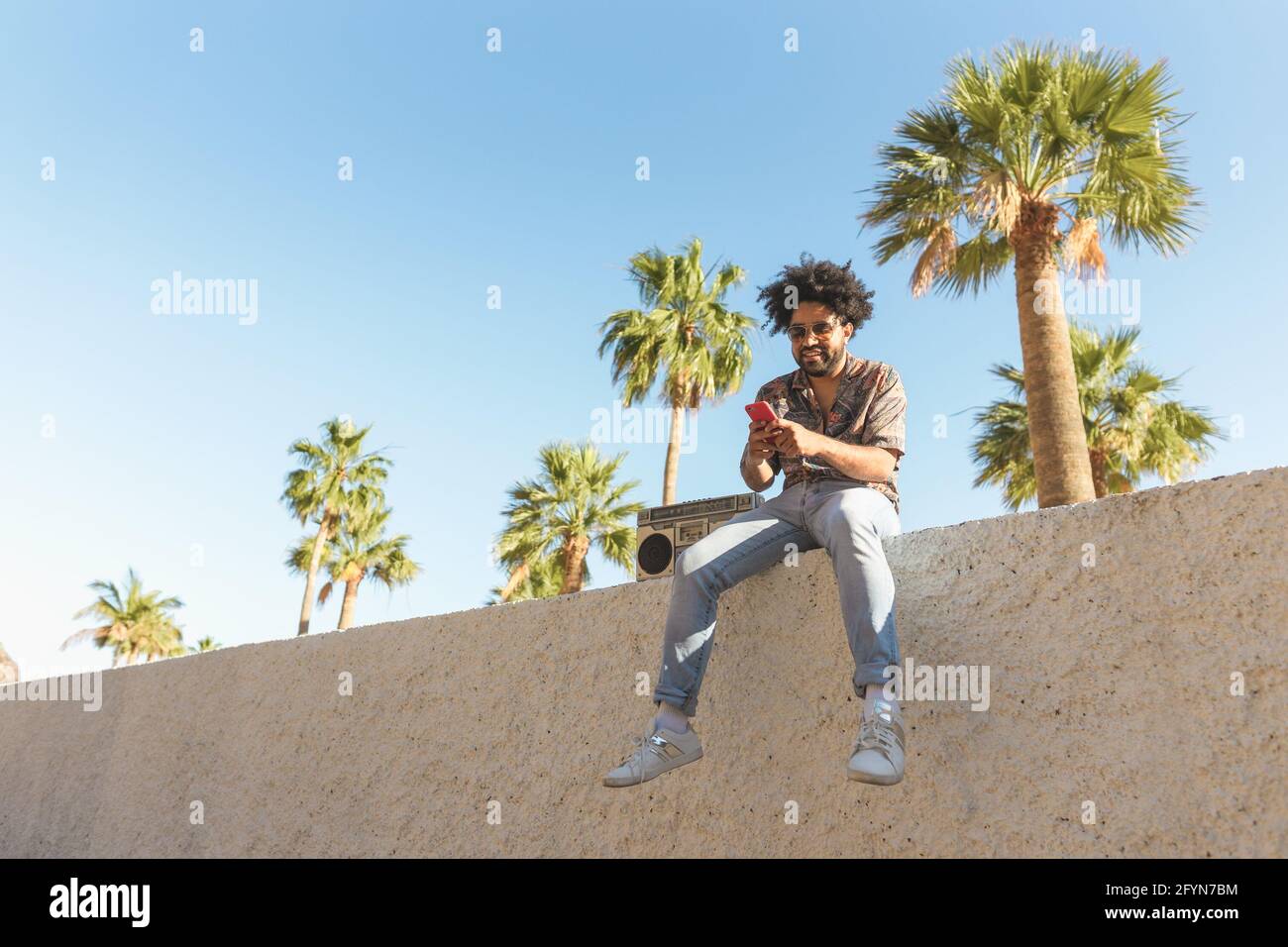 Joyeux Afro homme s'amusant à utiliser un smartphone mobile tout en écoutant à la musique avec stéréo boombox vintage dans un endroit tropical pendant vacances d'été Banque D'Images