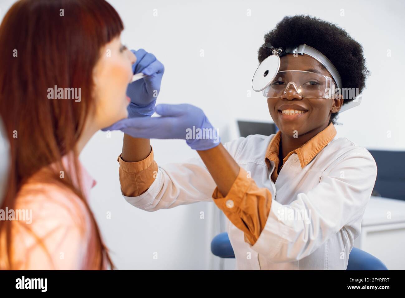 Positif sourire jeune Afro-américaine femme médecin otorhinolaryngologiste, portant uniforme, réflecteur frontal et lunettes, vérifiant la gorge de sa patiente, en utilisant dépresseur de langue Banque D'Images