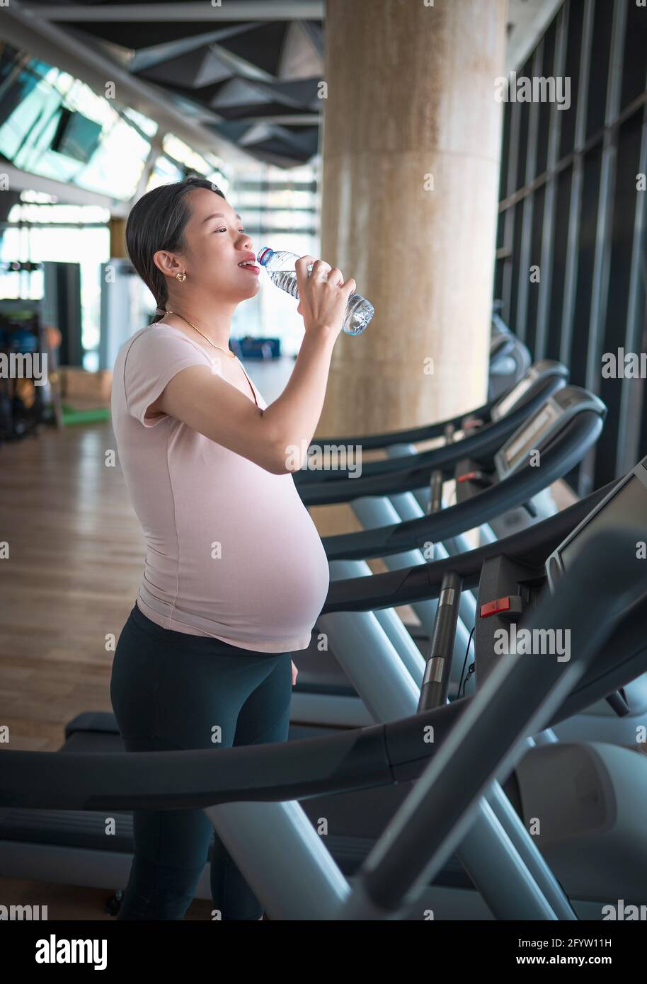 Femme asiatique enceinte hydratant pendant qu'elle fait de l'exercice dans la salle de sport à l'intérieur marcher sur un tapis roulant pour une forme et un style de vie actif pendant la grossesse Banque D'Images