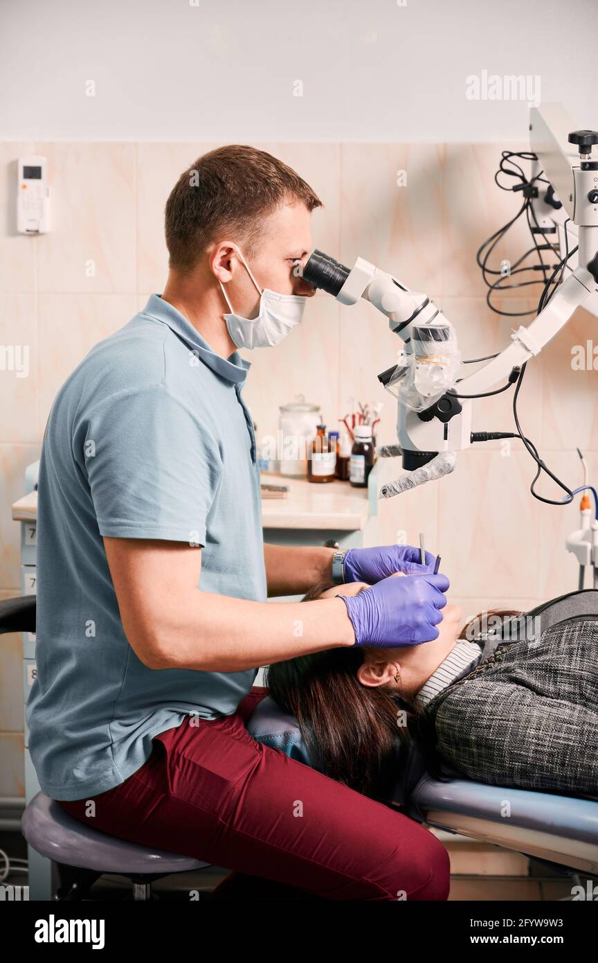 Dentiste de sexe masculin utilisant un microscope de diagnostic dentaire lors du traitement des dents du patient. Femme allongé sur une chaise dentaire pendant un examen dentaire dans un centre médical moderne. Concept de la dentisterie et des soins dentaires. Banque D'Images