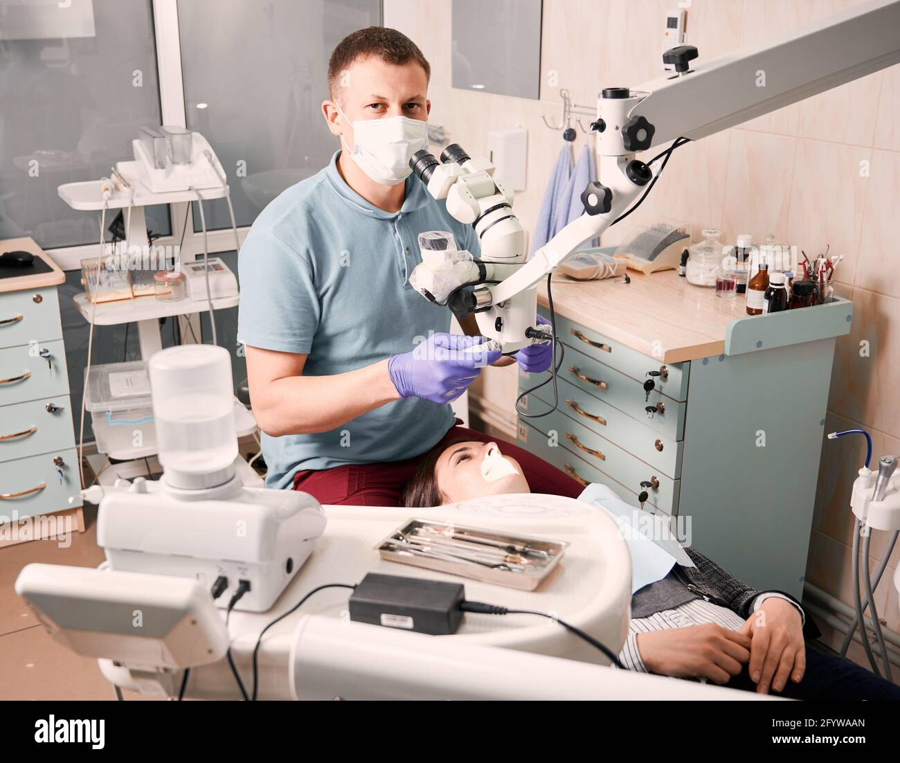 Jeune homme dentiste utilisant un microscope de diagnostic dentaire regardant la caméra pendant que la patiente est allongée dans une chaise dentaire. Concept de la dentisterie, de la stomatologie et des soins dentaires. Banque D'Images