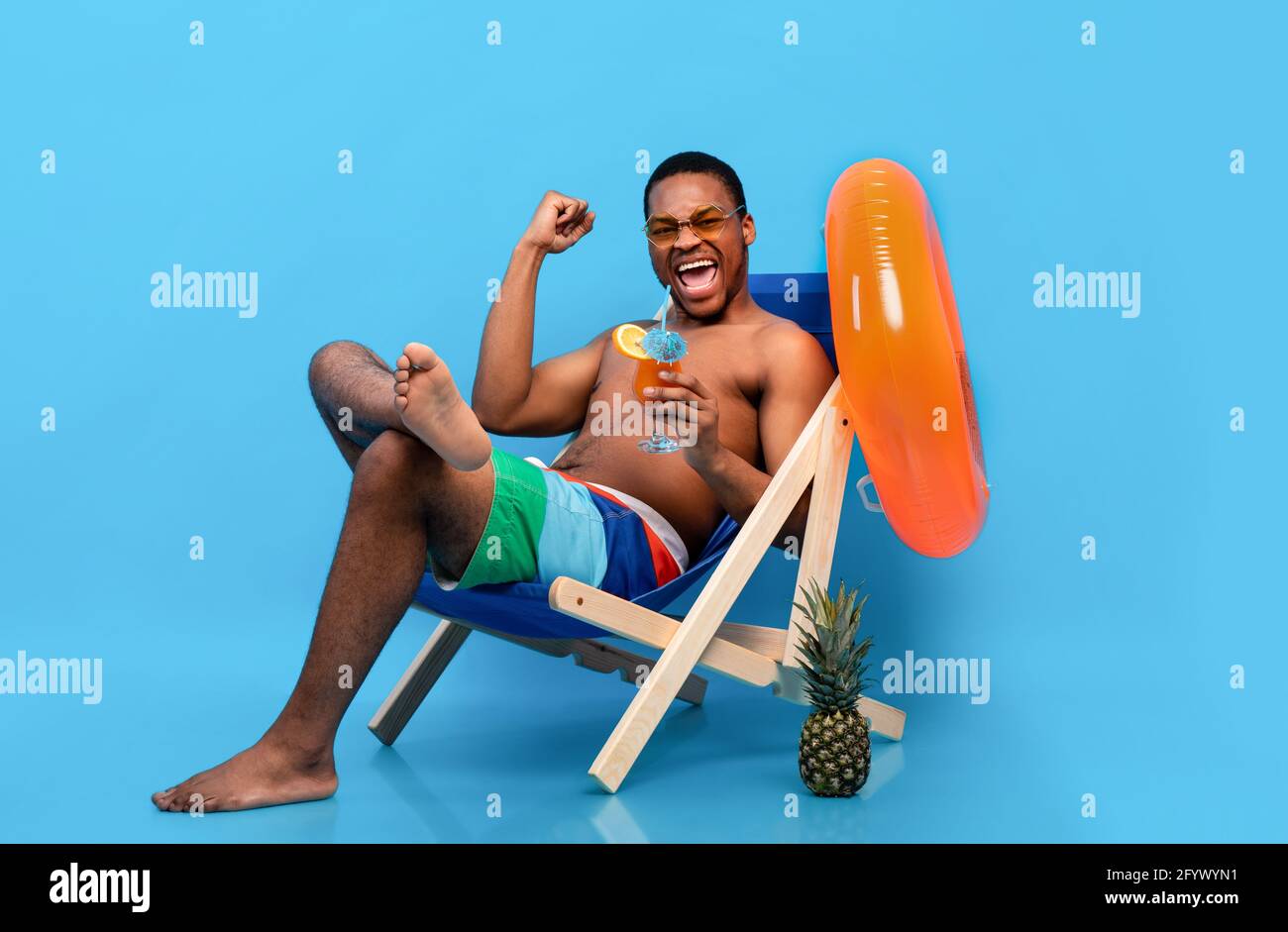 Un homme noir plein d'excitation qui boit un cocktail tropical dans une chaise longue, en faisant un geste sur fond bleu Banque D'Images
