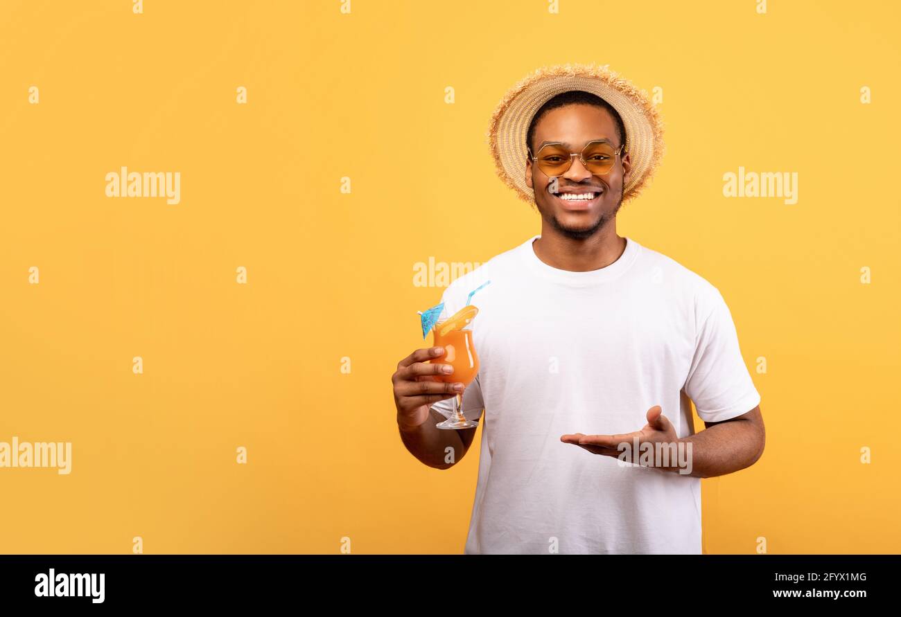 Happy Black Guy dans une tenue d'été offrant un verre de cocktail tropical sur fond jaune, bannière avec espace de copie Banque D'Images
