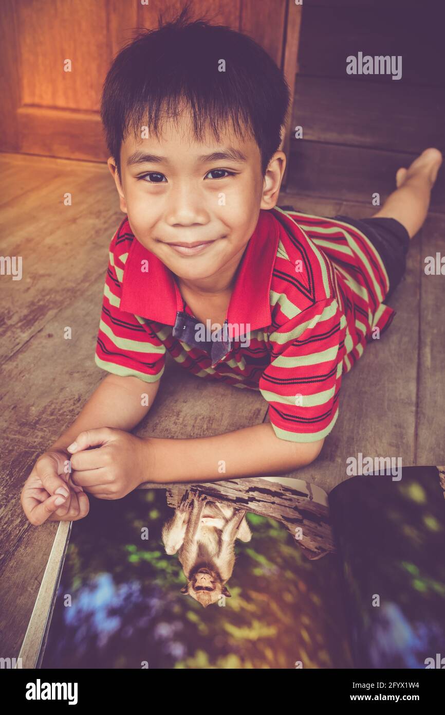 Beau garçon asiatique souriant et regardant l'appareil photo. Enfant lisant un album photo sur la faune animale, couché sur un sol en bois, à l'extérieur à la maison. Enfants Banque D'Images