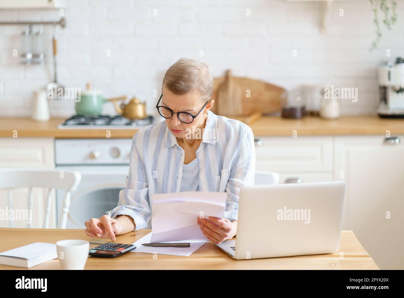 Femme comptable moderne, mature et concentrée, portant des lunettes pour analyser des documents, travaillant sur un ordinateur portable à domicile. Paiements par chèque de sexe féminin, compte sur la calculatrice. Concept senior et indépendant Banque D'Images
