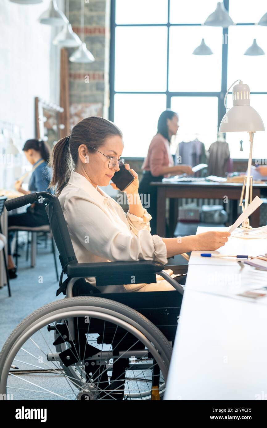 Femme en fauteuil roulant regardant à travers le papier et parlant au client sur un smartphone Banque D'Images