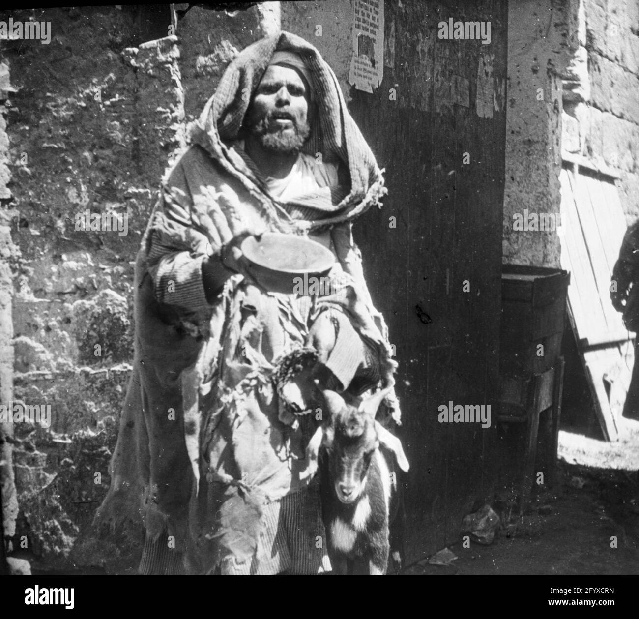 Un mendiant debout à côté d'un mur tient une corde attachée à une chèvre d'une main et un bol ouvert dans l'autre, Fès, Maroc, 1894. (Photo de Burton Holmes) Banque D'Images