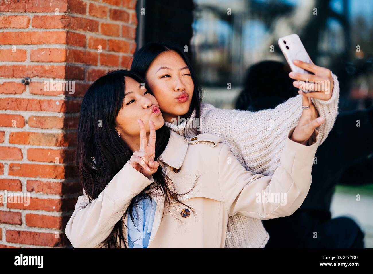 Des jeunes amies ethniques confiantes dans des vêtements élégants qui se replient sur les lèvres Et en affichant le signe V tout en prenant le selfie sur un téléphone portable debout dans la rue près de b Banque D'Images