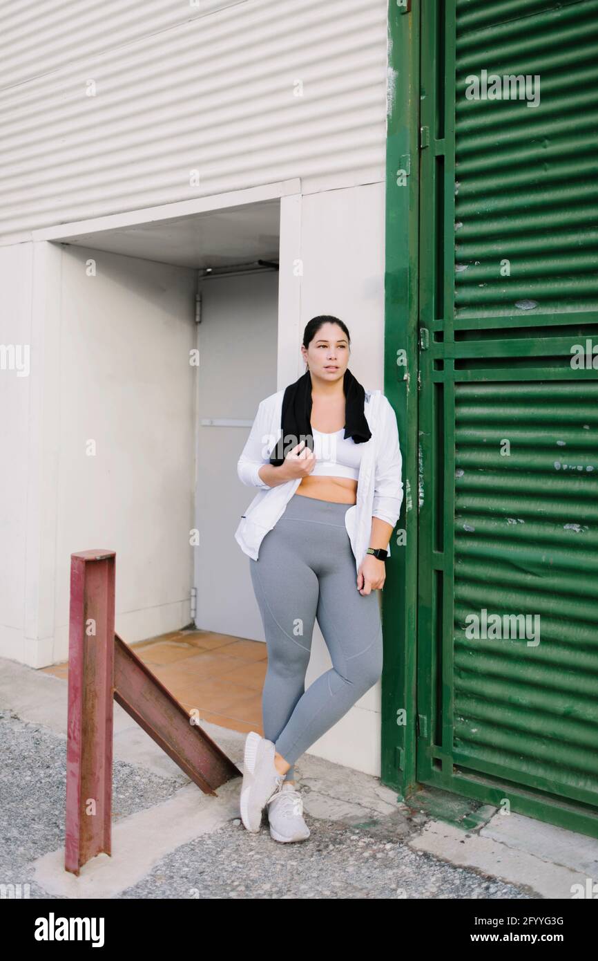 Pleine longueur de jeune athlète ethnique confiante dans les vêtements de sport s'appuyer sur la porte métallique d'un bâtiment vieillissant tout en se reposant entraînement en plein air Banque D'Images