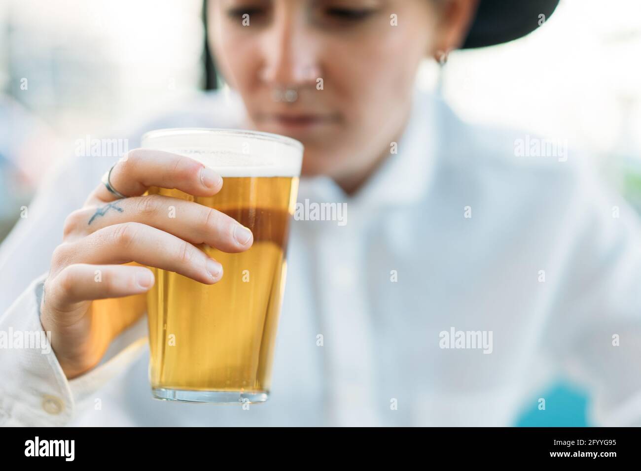 Petit garçon positif avec tatouages portant un chapeau et une chemise blanche boire de la bière du verre dans le café Banque D'Images