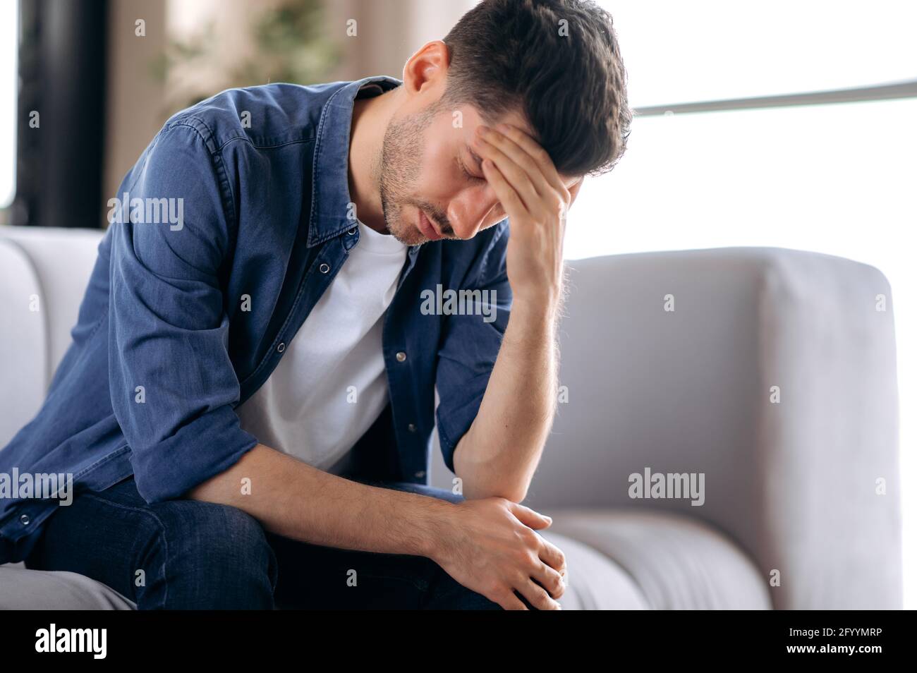 Bouleversé malade stressé jeune homme caucasien dans des vêtements décontractés assis sur le canapé dans le salon, se pencher, massant sa tête, souffrant de douleur, migraine, fermer les yeux, a besoin de traitement et de repos Banque D'Images