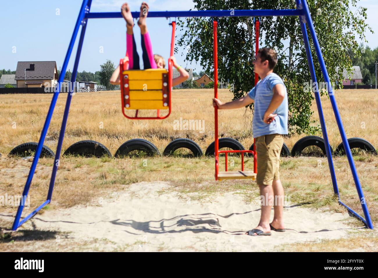 Recentrer la petite fille balançant sur l'oscillation sur le terrain de jeu avec le jeune homme, le gars, son frère plus âgé. Campagne. Swing bleu et rouge vif. Été en famille Banque D'Images