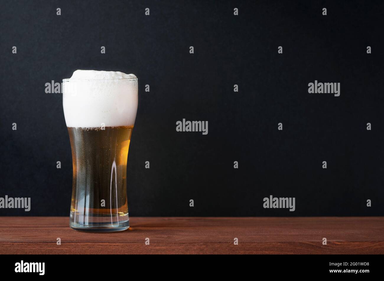 Bière froide mousseuse dans une tasse de bière en verre sur une table en bois dans un pub. Mur noir foncé comme arrière-plan pour le texte, le menu, la maquette Banque D'Images