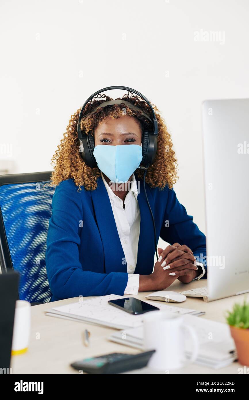 Portrait d'une jeune femme d'affaires gaie dans un casque et un masque médical salon au bureau devant l'ordinateur et vue à l'appareil photo Banque D'Images