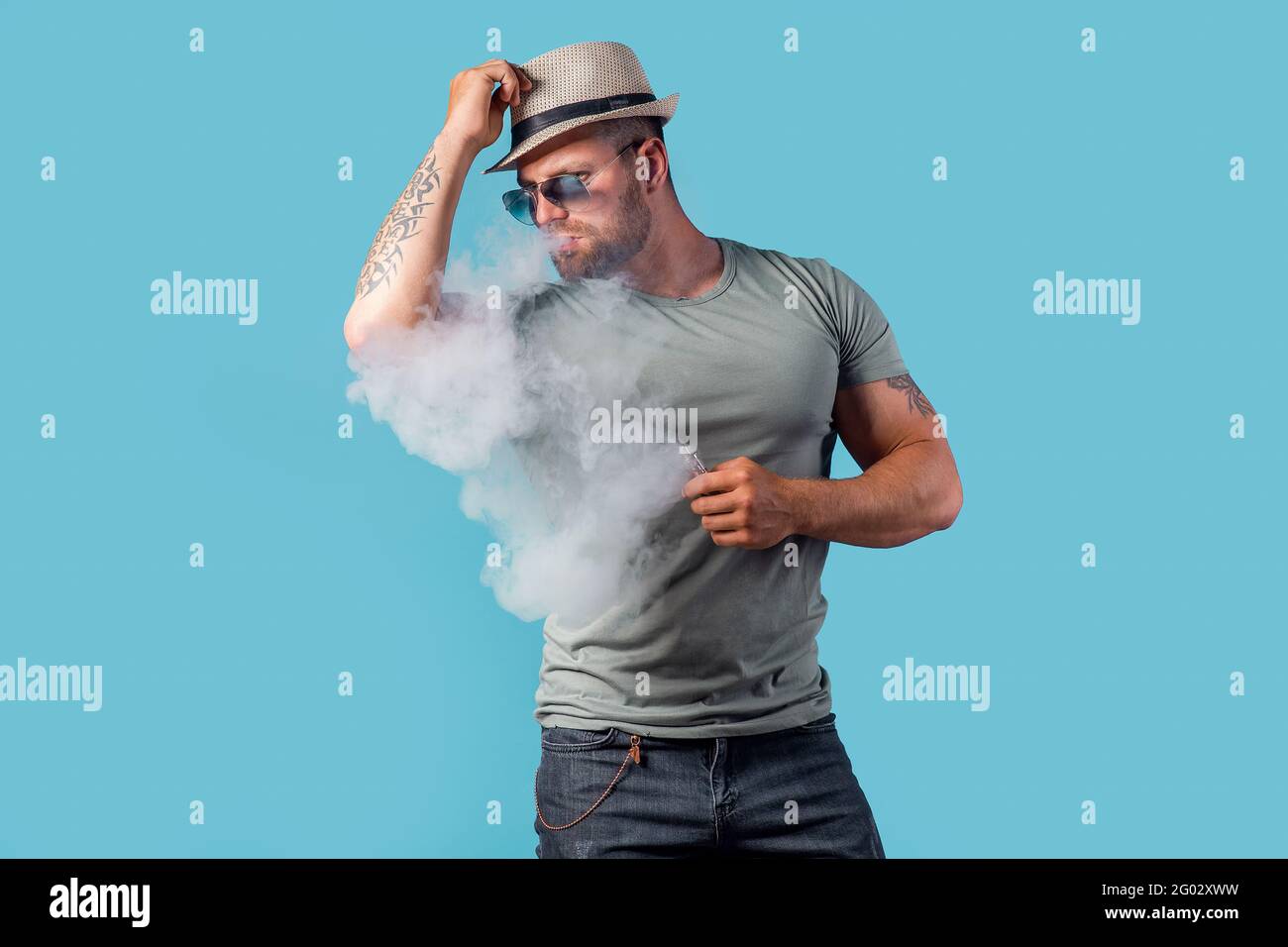 Mâle barbu en chapeau et lunettes de soleil fume une cigarette vapeur comme alternative au tabac. Studio tourné sur fond bleu. Banque D'Images
