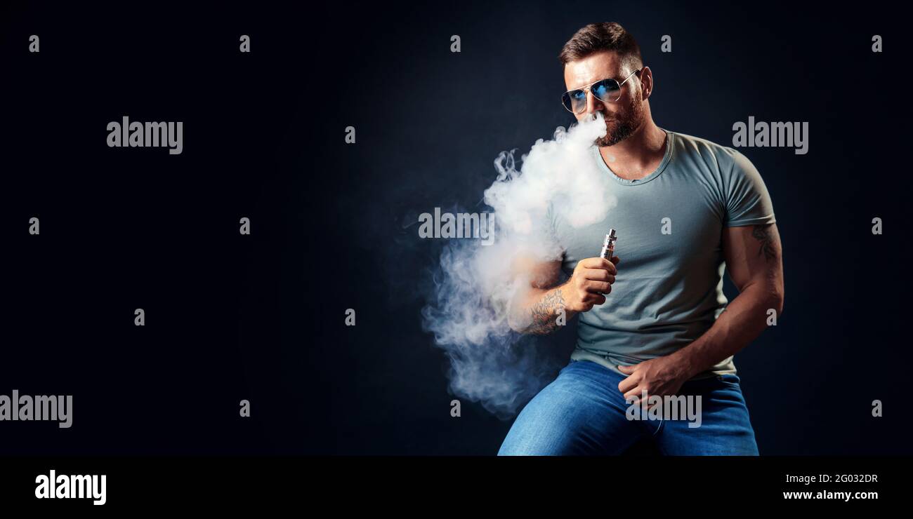 Mâle barbu brutal dans des lunettes de soleil fume une cigarette vapeur comme alternative au tabac. Studio tourné sur fond sombre. Banque D'Images