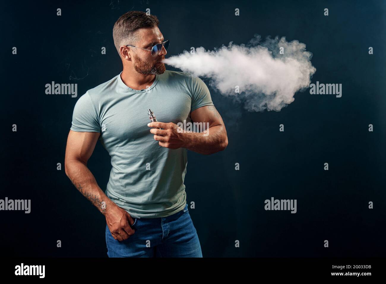 Mâle barbu brutal dans des lunettes de soleil fume une cigarette vapeur comme alternative au tabac. Studio tourné sur fond sombre. Banque D'Images