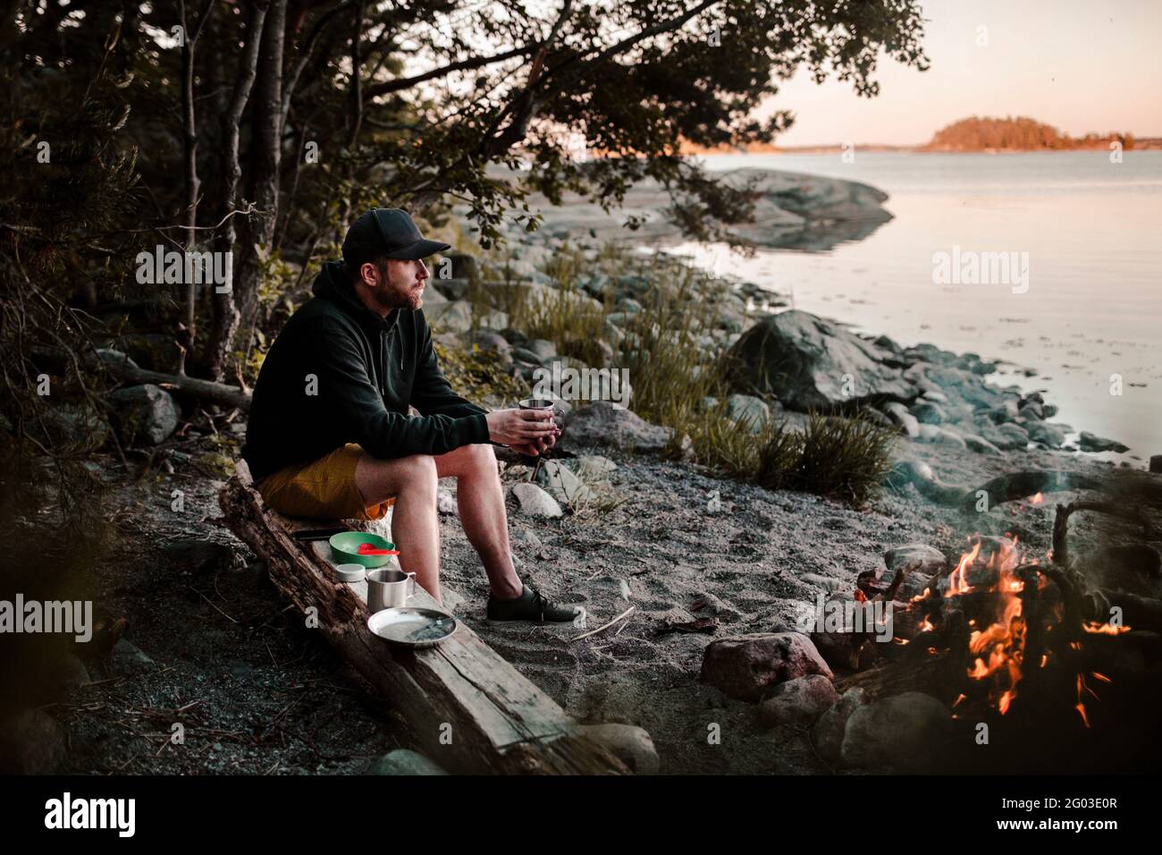 Pleine longueur d'homme regardant loin tout en étant assis sur une bûche près d'un feu de camp au bord du lac Banque D'Images