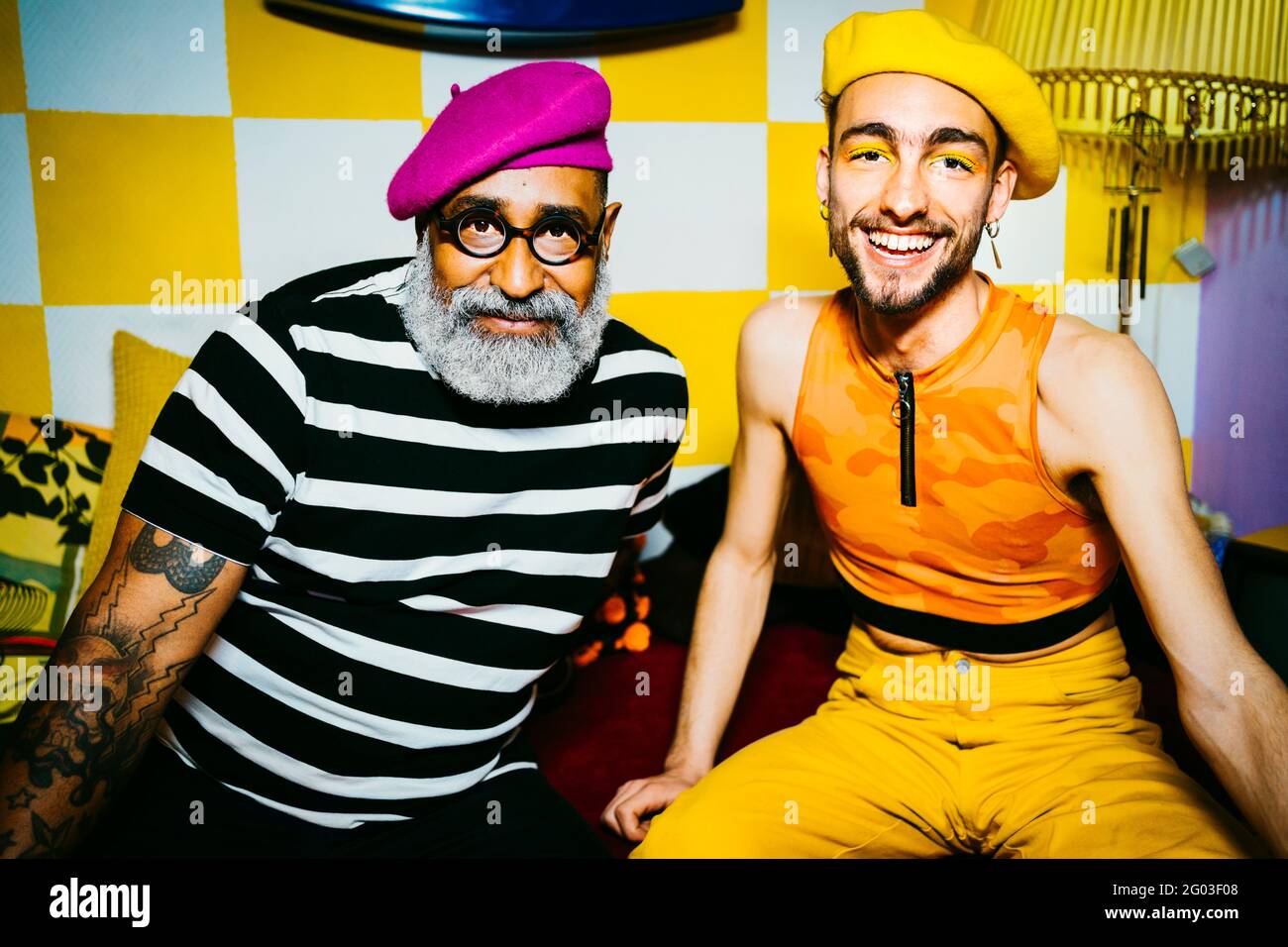 Jeune homme souriant avec un ami masculin senior portant des bérets assis dans la salle de séjour Banque D'Images