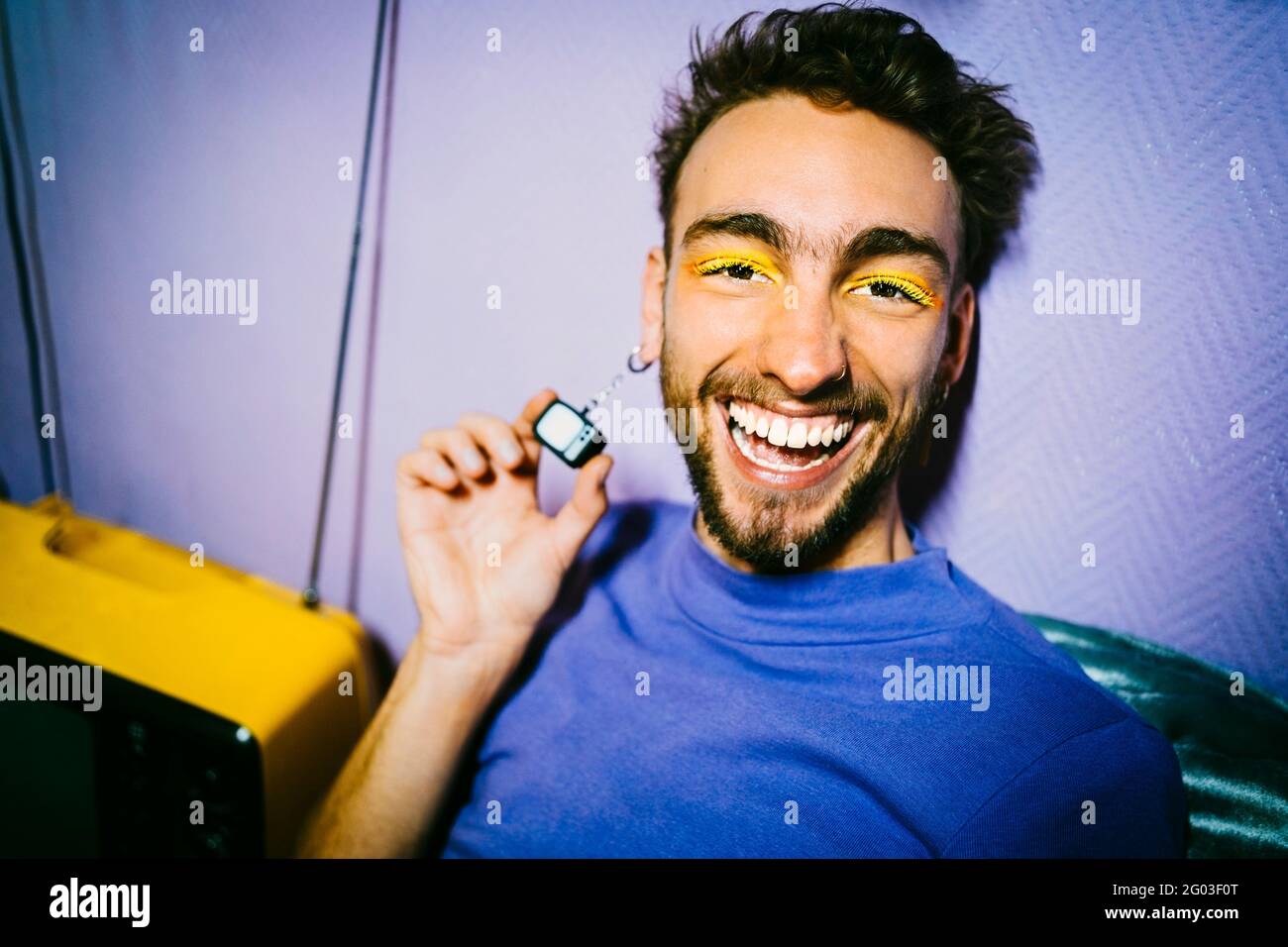 Portrait d'homme joyeux avec fard à paupières jaune montrant des boucles d'oreilles de télévision à l'appartement Banque D'Images