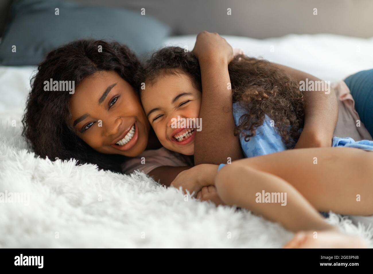 Parent amour enfant. Bonne jeune femme noire embrassant sa petite fille sur le lit, mignonne fille riant et appréciant la hug Banque D'Images