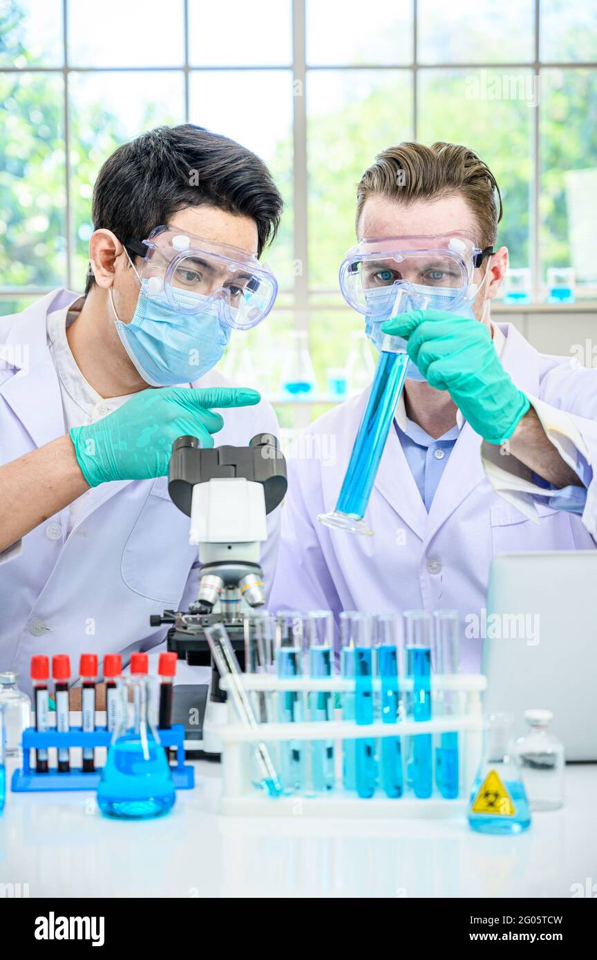 Couple scientifique mâle portant une combinaison de protection tenant le bécher ou le tube avec un échantillon de liquide à l'intérieur et en travaillant avec de nombreux équipements de laboratoire pour la recherche en laboratoire Banque D'Images