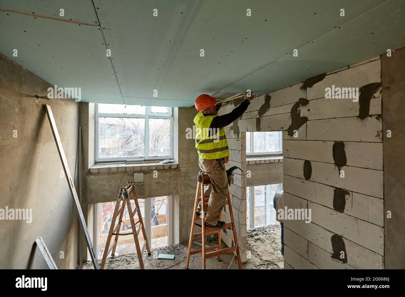 Travailleur professionnel debout sur une échelle et effectuant des mesures à l'aide d'un mètre ruban pour installer un plafond à cloisons sèches dans un bâtiment en hauteur en construction Banque D'Images