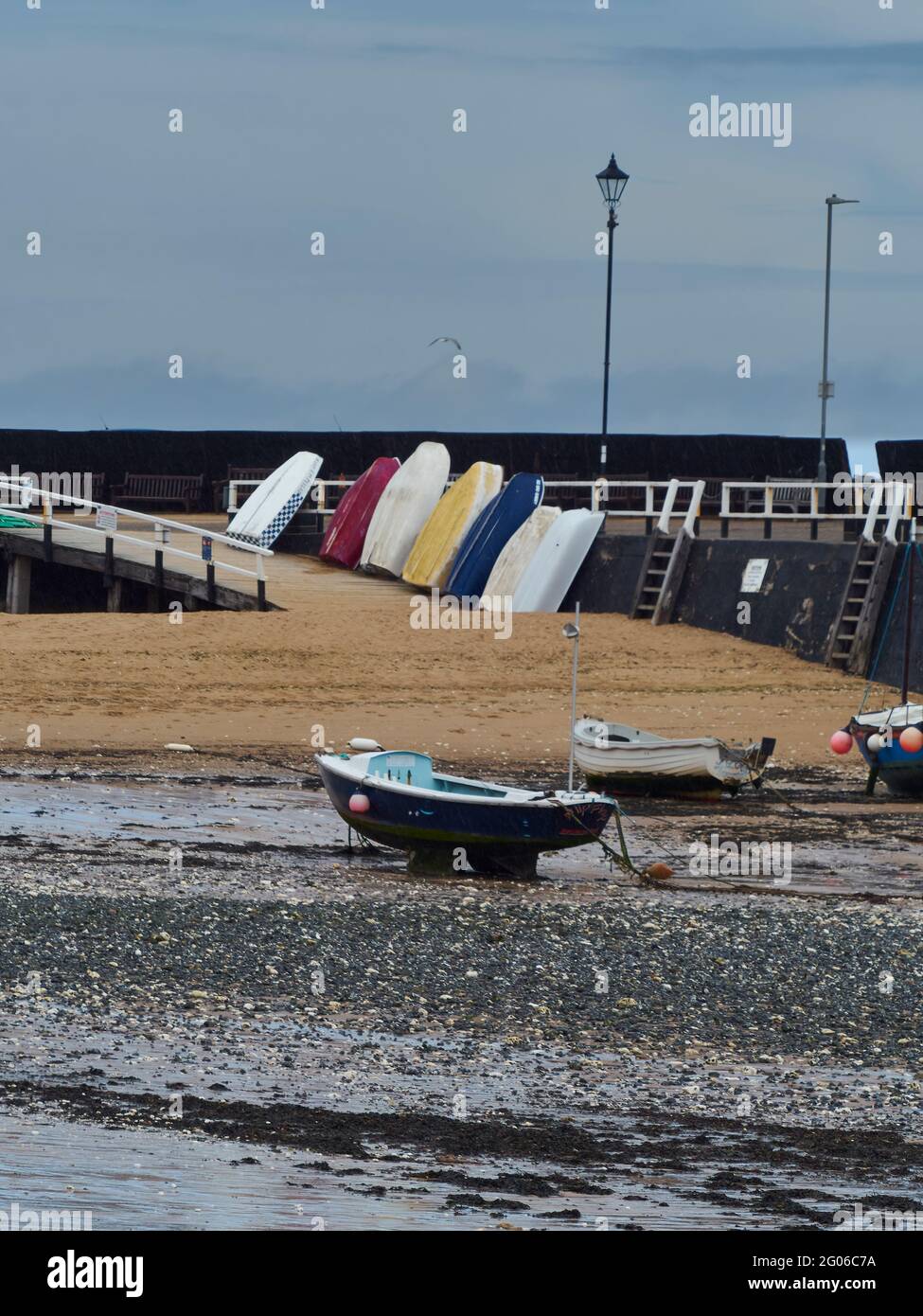 Petits bateaux au repos dans le sable et des galets derrière le brise-lames d'une ville de bord de mer, avec un magasin de gull à travers le fond. Banque D'Images