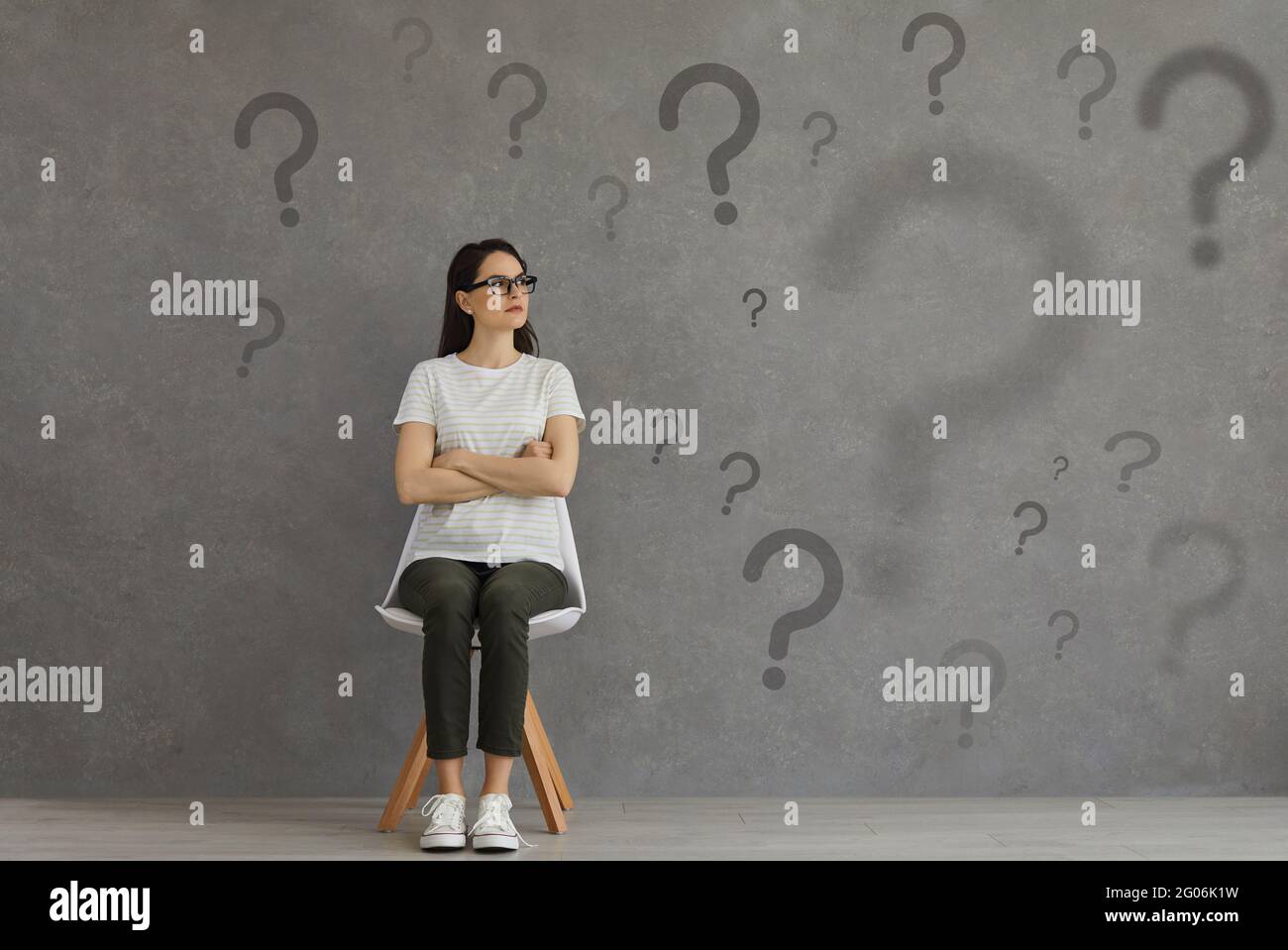 Femme confuse assise sur une chaise et regardant beaucoup de points d'interrogation sur fond gris Banque D'Images