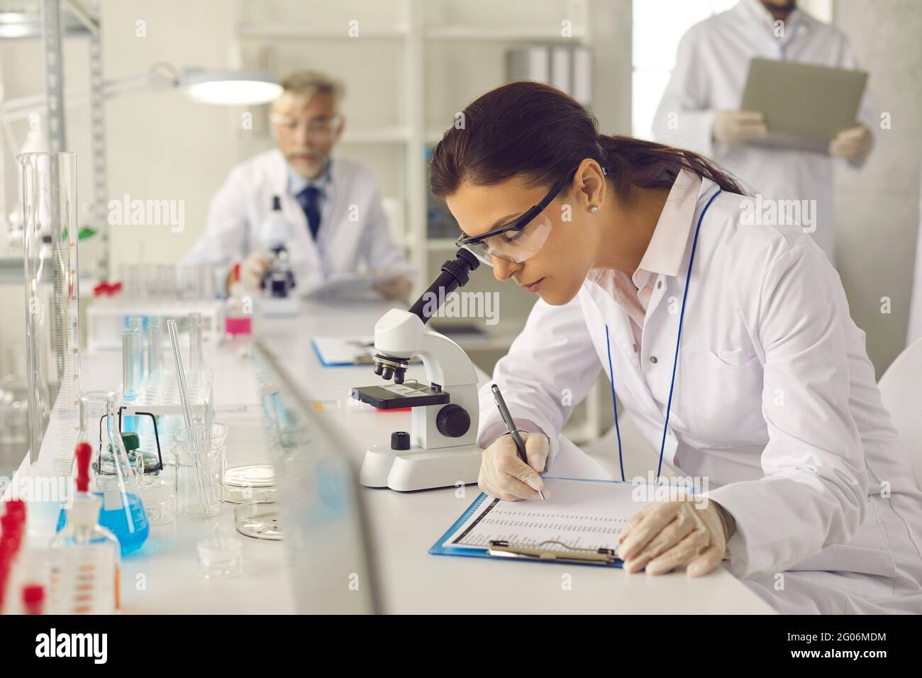 Scientifique en blouse de laboratoire, lunettes et gants regardant au microscope et prenant des notes Banque D'Images