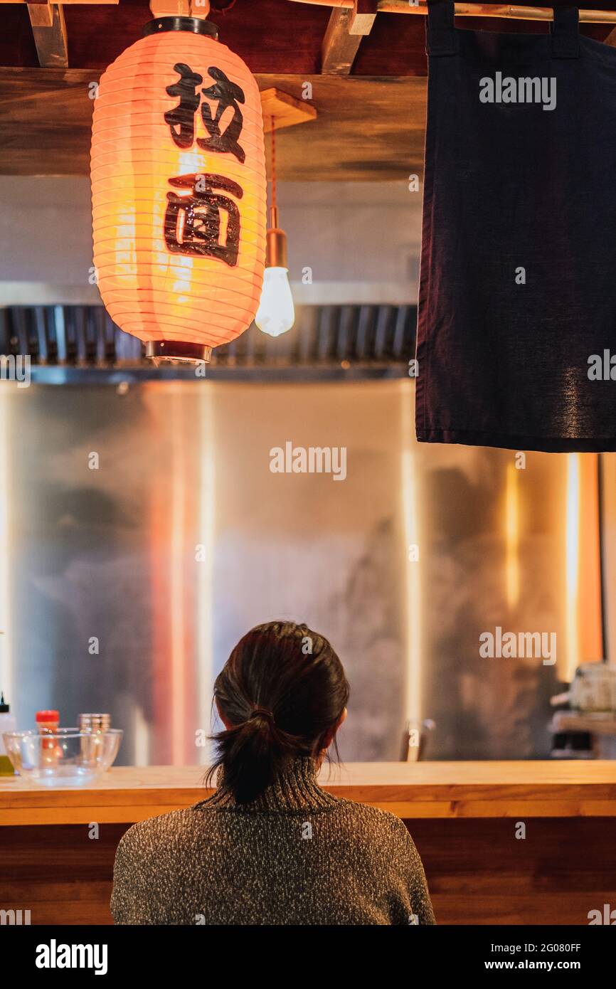 Vue arrière de la femme à cheveux noirs sans visage dans un chandail assis au comptoir dans un confortable bar ramen Banque D'Images